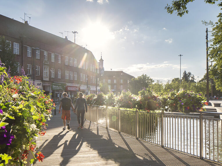 Two people walking down Watford Highlight Street. The sun is shining and there are flowers blooming.