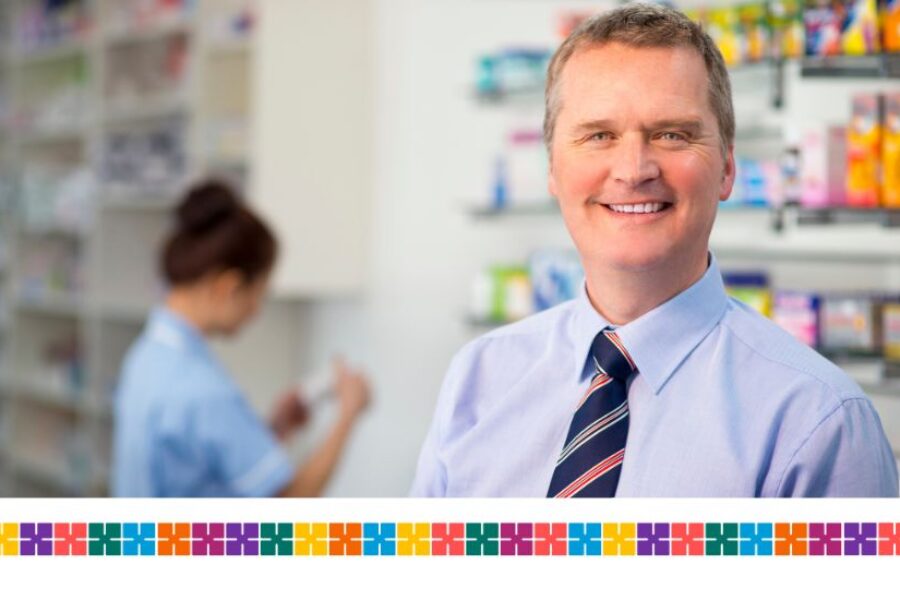 Male pharmacist smiling at camera. Pharmacy staff member looking at medicines behind him.